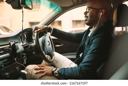 Side View Of Young Man In Suit Listening Music While Driving Car. African Businessman Driving A Vehicle To Office.