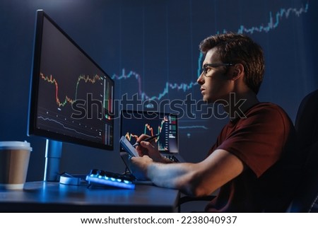 Side view of young man trader analyst at working table, analyzing financial chart, stock quotes, share prices, trading online, checking data on cryptocurrency graph on computer screen, making notes