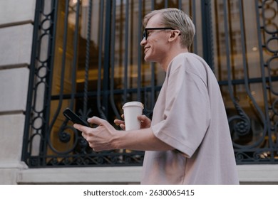 Side view of young man walking with coffee and phone, smiling while engaged with screen, captured against elegant European building with decorative iron bars