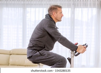 Side View Of Young Man Wearing Sportswear While Training On A Spin Bike In The Living Room. Shot At Home