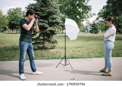 Side view of young man working with professional model. Photographer taking photos of pretty woman posing on street in park.