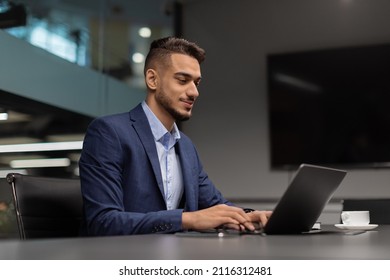 Side View Of Young Middle Eastern Businessman Sitting At Worktable At Modern Office, Typing On Notebook Keyboard, Sending Emails To His Business Partners, Working On Marketing Research, Copy Space