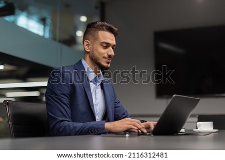 Side view of young middle eastern businessman sitting at worktable at modern office, typing on notebook keyboard, sending emails to his business partners, working on marketing research, copy space