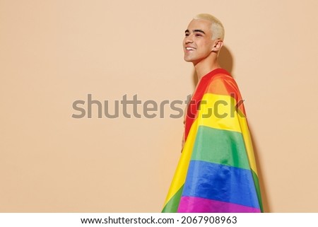 Side view young minded happy fun blond latin gay man with make up in beige tank shirt wrapped in rainbow flag look aside isolated on plain light ocher background studio People lgbt lifestyle concept