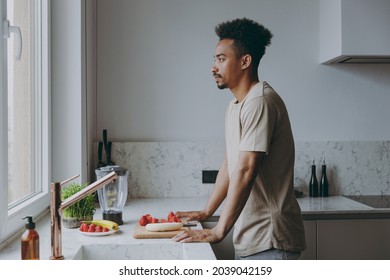 Side View Young Minded Serious African American Man 20s In Casual Clothes Look At Window Prepare Fruit Sweet Salad Cooking Food In Light Indoor Kitchen At Home Alone. Healthy Diet Lifestyle Concept