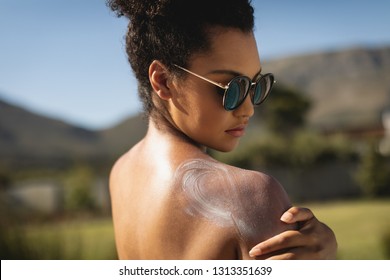 Side view of young mixed race woman applying sunscreen on shoulders in backyard of home on a sunny day