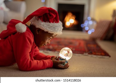 Side view of a young mixed race boy in his sitting room at Christmas, wearing a santa hat and holding a snow globe - Powered by Shutterstock