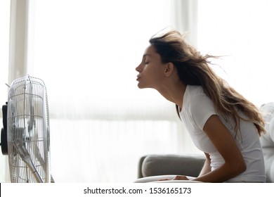 Side view young mixed race woman sitting on couch in front of ventilator indoors. Millennial girl suffering from hot summer weather or high temperature at home, cooling herself with air conditioner.