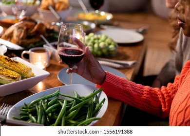 Side View Of A Young Mixed Race Woman Sitting At A Table At Home Set For Thanksgiving Dinner Holding A Glass Of Red Wine At The Table