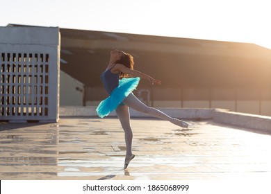 Side view of a young mixed race female ballet dancer wearing a blue tutu standing on one leg on her toes in a ballet pose, on the rooftop of an urban building, backlit by sunlight - Powered by Shutterstock