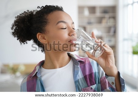 Side view of young mixed race girl drinking clean mineral still water from glass at home. Beautiful thirsty teen lady enjoying daily healthy lifestyle habit. Natural beauty, body skincare, wellness.