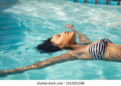 Side View Of Young Mixed-race Woman Floating In Swimming Pool In Backyard Of Her Home