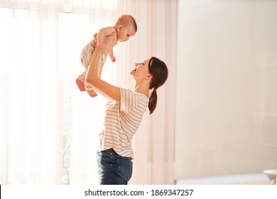 Side view of the young mom enjoying time with her newborn baby, while holding him in air and looking at him with pleasure while playing at home. Happy childhood concept