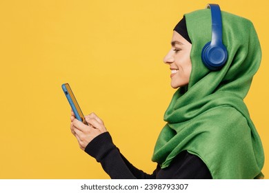 Side view young muslim woman wear green hijab abaya black clothes headphones listen music use mobile cell phone isolated on plain yellow background. People uae middle eastern islam religious concept - Powered by Shutterstock