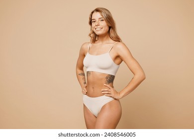 Side view young nice lady woman with slim body perfect skin wear nude top bra lingerie stand hold hands on waist look camera isolated on plain pastel light beige background. Lifestyle diet fit concept