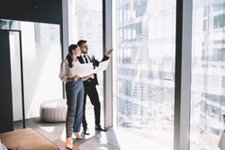 Side View Of Young Office Coworkers Standing Near Glass Wall In Modern Office And Looking Away At High Rise Buildings And Discussing With Documents In Hand
