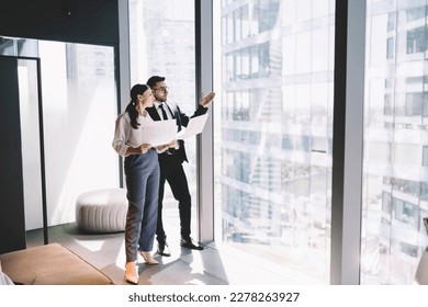Side view of young office coworkers standing near glass wall in modern office and looking away at high rise buildings and discussing with documents in hand