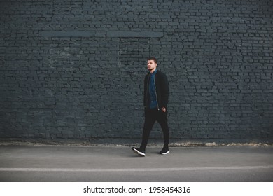 Side view of young pensive ethnic bearded man in trendy dark clothes walking along high gray brick wall on street