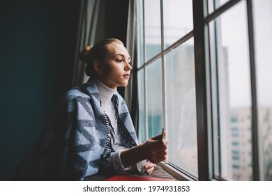 Side View Of Young Pensive Female Wrapping In Blanket Leaning On Windowsill While Looking Out Window Having Rest At Home