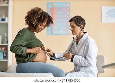 Side view of young pregnant African American woman looking at her big bare belly while senior Caucasian gynecologist checking baby with stethoscope in modern clinic - Powered by Shutterstock