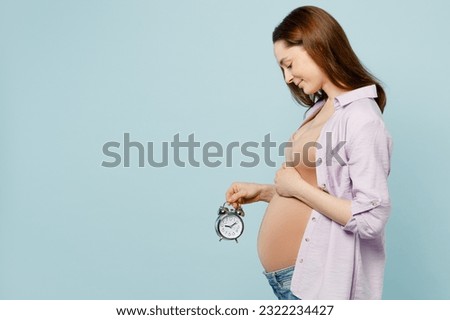 SIde view young pregnant future mom woman with belly tummy with baby wear casual clothes hold in hand clock alarm stroking stomach isolated on plain pastel blue background. Maternity pregnancy concept