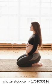 Side View Of Young Pregnant Woman Sits On Her Knees On Mat Doing Hip Stretching Exercises, Hugs Her Tummy, Looks In Front Of Her. Vertical, Full Length, Copy Space, Pregnancy Yoga And Fitness Concept