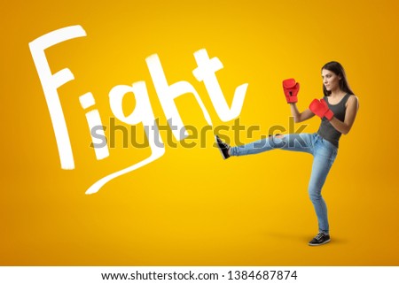 Side view of young pretty girl in jeans, sleeveless top and red boxing gloves with leg lifted for a kick on yellow background with Fight title. Show fighting spirit. Never give up. Heated rivalry.