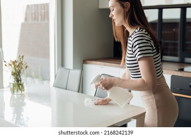 Side View Of Young Pretty Woman Standing In Kitchen, Spraying On White Table With Vase From Pulverizer, Wiping With Rag.