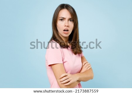 Side view young sad indigant disturbed woman 20s wearing pink t-shirt look camera hold hands crossed folded isolated on pastel plain light blue background studio portrait. People lifestyle concept.