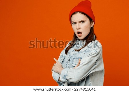Side view young sad mad furious angry indignant woman wear denim shirt white t-shirt red hat hold hands crossed folded look camera isolated on plain orange background studio portrait Lifestyle concept