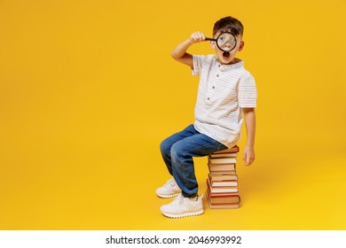 Side View Young School Boy 5-6 Years Old In Casual Clothes Sit On Pile Of Books Looking Through Magnifier Isolated On Plain Yellow Background Studio Childhood Children Kids Education Lifestyle Concept