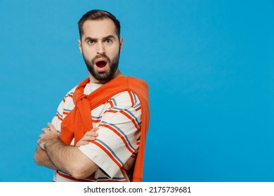 SIde view young serious dissatisfied displeased indignant man 20s in orange striped t-shirt looking camera hold hands crossed folded isolated on plain blue background studio. People lifestyle concept