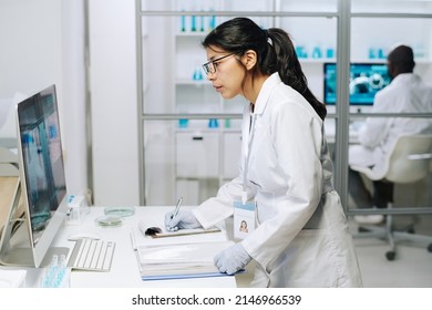 Side View Of Young Serious Female Clinician Looking At Computer Screen With Coronavirus Macro Image While Making Notes In Document