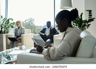 Side View Of Young Serious Female Office Worker With Tablet Sitting On White Leather Couch In Openspace Office Against Two Coworkers