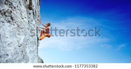 side view of young slim woman rock climber in bright orange pants climbing on the cliff. a woman climbs on a vertical rock wall on the blue sky background