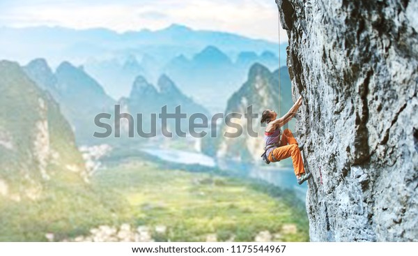 side view of young slim woman rock climber in\
bright orange pants climbing on the cliff against a high mountains.\
girl climbs on a vertical flat rocky wall and making hard move.\
Copy space
