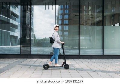 Vista lateral de una joven mujer de negocios sonriente con casco de bicicleta en la cabeza conduciendo una motocicleta eléctrica