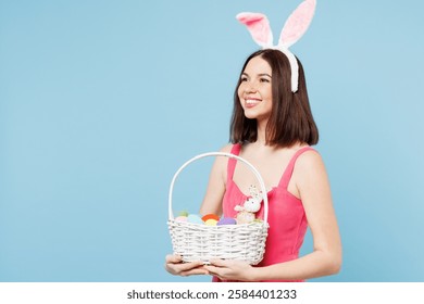 Side view young smiling fun woman wears pink casual clothes rabbit bunny ears hold wicker basket with eggs look aside isolated on plain blue background studio portrait. Lifestyle Happy Easter concept