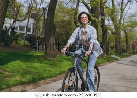 Side view young smiling fun sporty woman in jeans clothes headphones riding bicycle bike on sidewalk in city spring park outdoors, look aside. People active urban healthy lifestyle cycling concept.