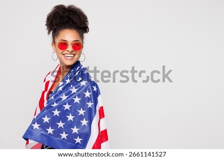 Side view young smiling fun woman of African American ethnicity wrapped in American flag wear sunglasses look camera isolated on plain solid white background studio. Lifestyle, the 4th of July concept