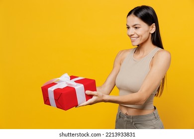 Side View Young Smiling Happy Cheerful Cun Cool Latin Woman 30s She Wear Basic Beige Tank Shirt Hold In Hand Give Red Present Box With Gift Ribbon Bow Isolated On Plain Yellow Color Backround Studio