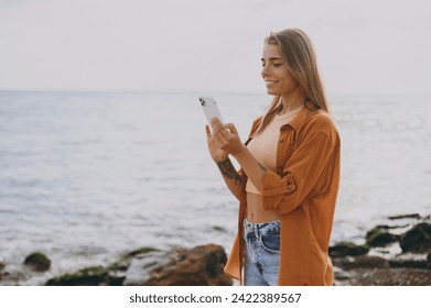 Side view young smiling satisfied woman she wearing orange shirt casual clothes hold in hand use mobile cell phone walk on sea ocean sand shore beach outdoor seaside in summer day. Lifestyle concept - Powered by Shutterstock