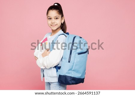 Side view young smiling smart school girl 8-9 years old wearing blue casual clothes backpack hold books isolated on plain pink background children portrait. Childhood kids education lifestyle concept