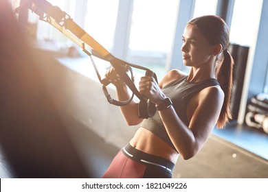Side view of a young strong and fit woman exercising with fitness trx straps at gym