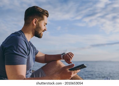Side view young strong sporty fit fun sportsman man in sports clothes shorts warm up train use mobile cell phone smart watch at sunrise sun over sea beach outdoor on pier seaside in summer day morning
