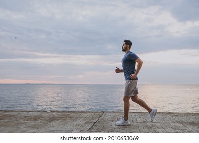 Side View Young Strong Sporty Athletic Toned Fit Sportsman Man 20s In Sports Clothes Warm Up Training Run Jogging At Sunrise Sun Over Sea Beach Outdoor On Pier Seaside In Summer Day Cloudy Morning.