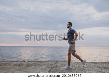 Side view young strong sporty athletic toned fit sportsman man 20s in sports clothes warm up training run jogging at sunrise sun over sea beach outdoor on pier seaside in summer day cloudy morning.