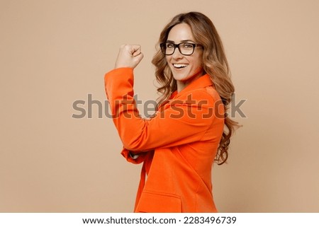 Side view young strong sporty fitness employee business woman corporate lawyer wear classic formal orange suit glasses work in office showing biceps muscles on hand isolated on plain beige background