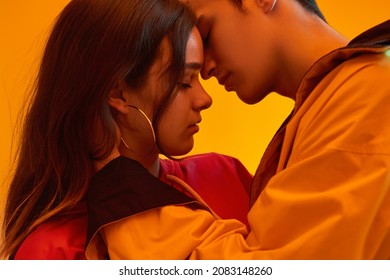 Side view of young trendy couple gently touching faces with closed eyes on orange background in studio