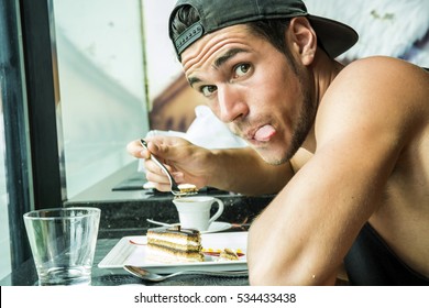 Side View Of Young Trendy Man Eating Dessert In Cafe And Showing Tongue Out At Camera. 